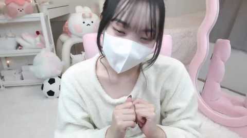 Media: Video of an East Asian woman with black hair, wearing a white face mask, pink sweater, and a pink chair, surrounded by plush toys in a pastel-themed room.