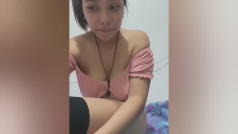 Media: Video of an Asian woman with dark hair, wearing a pink off-shoulder top, sitting in a white room, partially obscured by a black stocking.