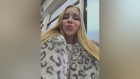 A video of a young, blonde Caucasian woman with long, straight hair, wearing a leopard-print jacket, making a playful face. The background features a window and indistinct indoor setting.