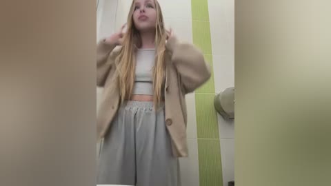 A video of a young blonde woman in a beige cardigan and grey sweatpants, standing in a tiled bathroom with green and white tiles, adjusting her hair.