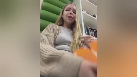 A video of a blonde woman with long hair, wearing a beige cardigan over a gray top, sipping from an orange cup, seated in a green chair against a bookshelf backdrop.