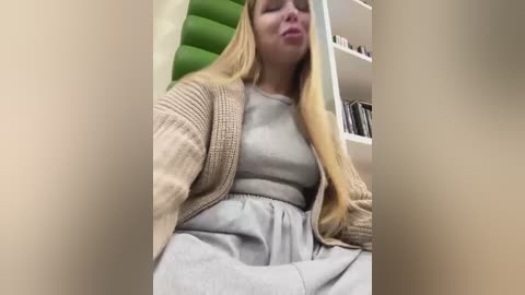 A video of a blonde woman with a round face, wearing a beige cardigan over a light grey dress, sitting on a couch in a room with a green wall and bookshelves.