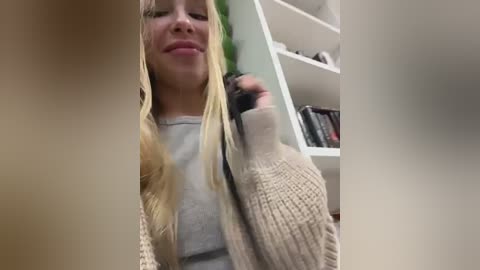 Video of a blonde woman with long hair, wearing a gray sweater and beige cardigan, smiling, holding a black object in a bookshelf-filled room.