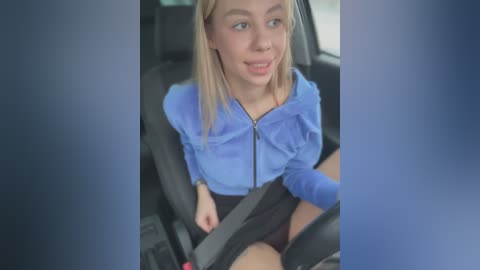 Media: Video of a young, light-skinned woman with straight blonde hair, wearing a blue hoodie and black shorts, sitting in a car seat, smiling.