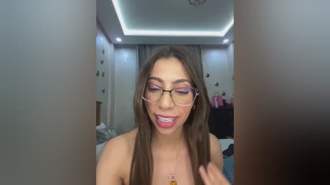 Media: Video of a young woman with long brown hair, wearing glasses and a necklace, smiling with braces. She stands in a brightly lit bedroom with white walls, a bed, and a ceiling light.