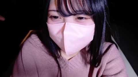 Media: Video of an Asian woman with long black hair, wearing a pink face mask, pink sweater, and a dark background.