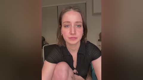 Video of a young Caucasian woman with light skin, shoulder-length brown hair, and blue eyes, wearing a black mesh top, sitting on a couch in a dimly lit room with beige walls.