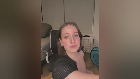 Media: Video of a young woman with shoulder-length brown hair, wearing a black mesh top, sitting on a gaming chair in a cluttered living room with beige walls, wooden floor, and a white laundry basket.