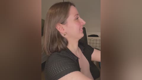 A video of a smiling Caucasian woman with shoulder-length brown hair, wearing a black textured top and a necklace, looking to the right in a dimly lit room.