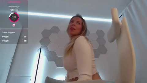 Media: Video of a white woman with long blonde hair, wearing a white sweater, standing in a modern, white room with hexagonal tiles on the wall.