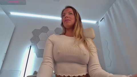 Media: Video of a blonde, light-skinned woman in a ribbed, white sweater, sitting on a beige chair, surrounded by modern, white decor with hexagonal wall panels and a floor lamp.