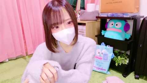 Video of an Asian woman with short brown hair, wearing a white mask and gray sweater, sitting on a green carpeted floor. Background features pink curtains, a black shelf with a blue plush toy, and a box.