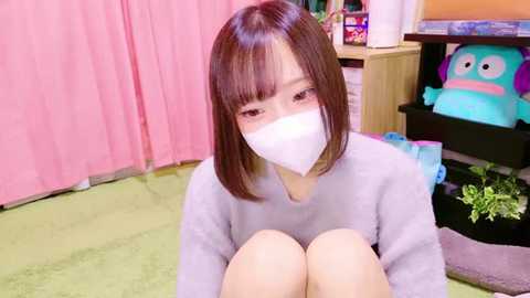 Media: Video of an East Asian woman with straight brown hair and bangs, wearing a white face mask and gray sweater, seated on a light green carpet. Background features pink curtains, a toy box with plush toys, and a plant.