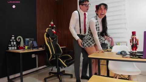 Media: Video of a man in glasses and a red tie, wearing a white shirt and suspenders, standing behind a woman in a plaid skirt and white top, in a modern office with wooden walls, desks, and chairs.