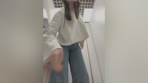 Media: Video of a woman with light skin, long brown hair, wearing a cream-colored sweater and high-waisted, loose-fitting jeans, standing in a narrow, beige hallway with white ceiling tiles.