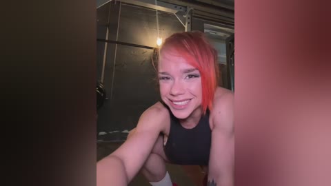 Media: Video of a smiling, athletic young woman with bright orange hair, wearing a black sports bra, crouching in a dimly lit, industrial space with exposed pipes and a metal wall.