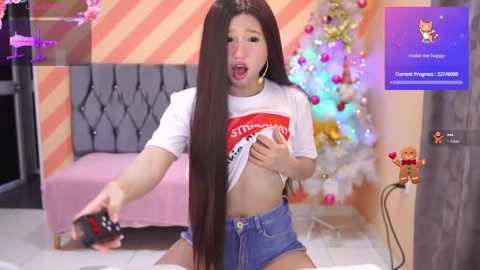 Media: Video of a young Asian woman in a white T-shirt, blue shorts, and hoop earrings, playfully lifting her shirt to reveal her midriff, standing in a festive, colorful room with a Christmas tree and toys.