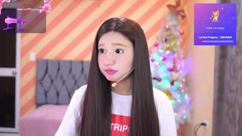 Media: Video of a young woman with long black hair, wearing a headset, white T-shirt, and pink lipstick, standing in a room with a striped wall, Christmas tree, and \"Make a Wish\" sign.