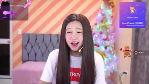 Media: Video of a young Asian girl with long black hair, wearing a white shirt, in a cozy, festive room with a decorated Christmas tree, pink bed, and a pink wall.