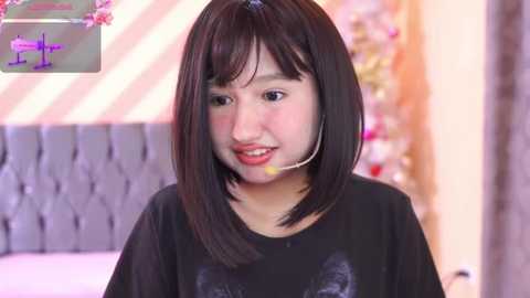 Media: Video of a young Asian woman with straight, shoulder-length black hair, wearing a black T-shirt, a headset with a microphone, and a pink flower headband. Background features a pink and white striped wall and a grey cushioned headboard.