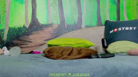 Media: A video of a messy bedroom with a green wall mural of a forest, a gray bed, and scattered clothes, including a pink bra and a brown blanket.