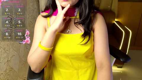 Media: Video of a woman with long black hair, wearing a bright yellow sleeveless dress and pink scarf, seated in a modern, dimly lit room with a digital clock and wooden decor.