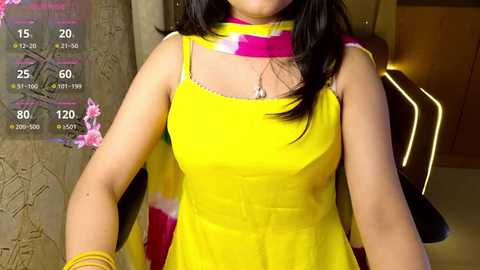 Media: Video of a woman wearing a bright yellow sleeveless dress with a pink and white scarf, in an indoor setting with a textured wall and modern lighting.