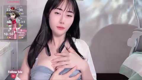 Media: Video of a young East Asian woman with long black hair, wearing a white cardigan and blue bra, sitting on a bed in a dimly lit room. The image has a \"Follow Me\" overlay.