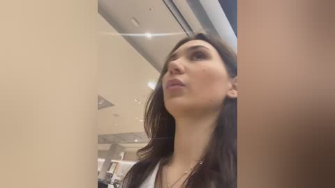 Media: Video of a light-skinned woman with long dark hair, wearing a white shirt and black jacket, looking up with a pensive expression, in a modern, well-lit room with beige walls and metallic accents.