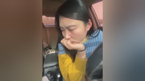 Media: Video of an Asian woman with long black hair, wearing a yellow sweater and blue blouse, resting her head on her hand while seated in a car.