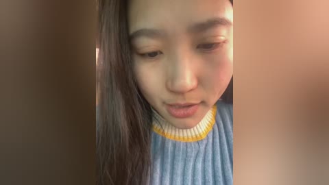 Media: A close-up video of a young Asian woman with long dark hair, wearing a light blue sweater with yellow trim, looking down with a slight frown. The background is blurred.