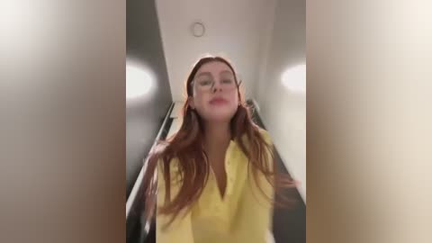 Media: A video captures a woman with long, wavy red hair, wearing a yellow blouse, standing in a dimly lit, narrow hallway with recessed lighting.