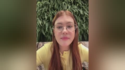 Media: Video of a young woman with long, straight red hair, fair skin, and large, round glasses, wearing a yellow shirt, seated in front of a lush green fern wall.