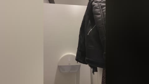 Media: A video of a modern, minimalist restroom interior featuring a black leather jacket hanging on a white wall, adjacent to a white urinal. The room has a clean, utilitarian design with no visible decorations.