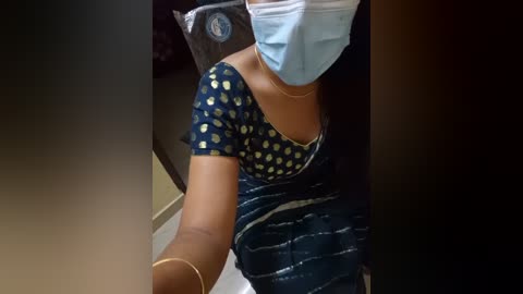 Media: A video of a woman with dark skin, wearing a blue face mask, dark blue top with white polka dots, and a gold bracelet, taking a selfie in a dimly lit room.