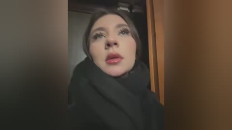 Media: A video of a young woman with fair skin, brown hair, and full lips, wearing a black scarf and coat, peering through a doorway. The background is dimly lit, with wooden and dark walls.