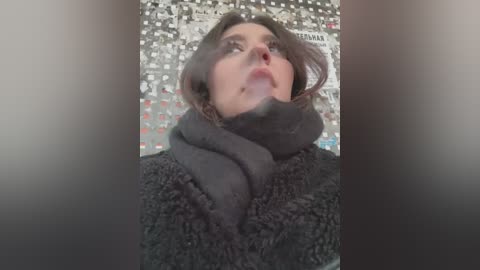 Media: Video of a woman with light skin and brown hair, wearing a dark, textured scarf and a fuzzy black sweater, against a mosaic tile background.