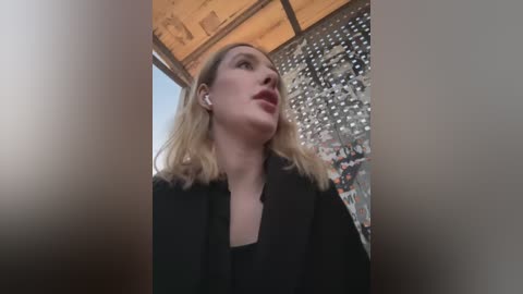 Media: Video of a blonde woman with fair skin, wearing a black blazer, looking up, surrounded by wooden beams and a metallic, reflective surface.