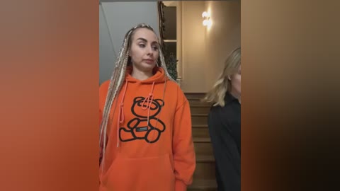 Media: Video of a young woman with light skin, long blonde dreadlocks, wearing an orange hoodie with a teddy bear graphic, walking indoors.