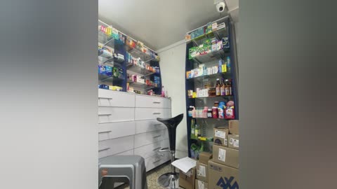 A video of a compact, organized pharmacy with white shelves filled with colorful medicines, a black stool, and stacked brown boxes on the floor.