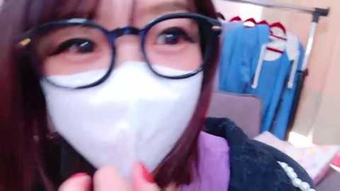 Media: A video of a young woman with long, straight dark hair, wearing glasses, and a white mask covering her nose and mouth, standing in a bright, organized room with blue clothing hanging on a rack in the background.