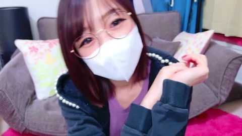 Video of an Asian woman with straight, shoulder-length dark hair, wearing glasses, a white face mask, and a purple top. She's holding a red pen in a living room with a grey couch and colorful pillows.