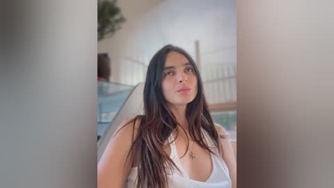 Media: Video of a young woman with long, straight black hair, wearing a white tank top, standing indoors near a staircase with a potted plant in the background.