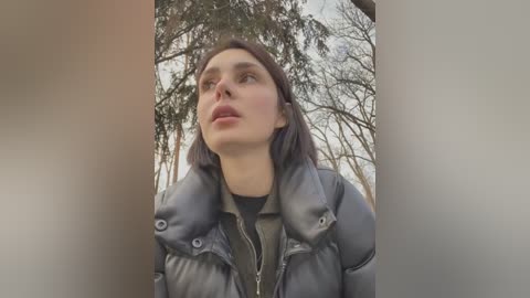 Media: A video of a young woman with fair skin, brown hair, and a grey puffy jacket, standing outdoors with bare trees in the background, looking upward.