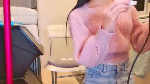 Media: Video of an East Asian woman with long black hair, wearing a pink knit sweater and high-waisted light blue jeans, standing in a bathroom with a white toilet, towel rack, and pink mirror.