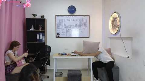 Media: Video of a minimalist office with white walls, a clock, shelves, and a white desk; a woman in a white top and black pants sits at the desk, while another woman in a pink dress and a man in a white shirt and black pants sit in chairs.