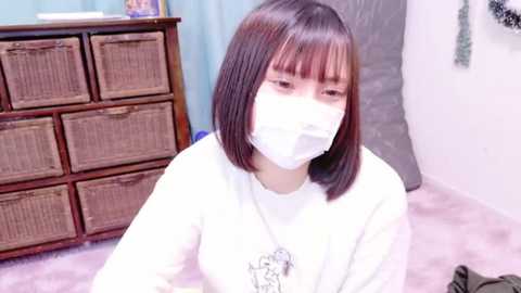 Media: Video of an Asian woman with straight, shoulder-length brown hair, wearing a white mask and shirt, seated on a pink carpet. Background features a wicker chest of drawers and a grey plastic bag.