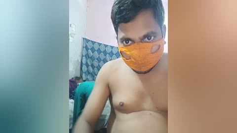 Media: Video of a shirtless man with medium build, light brown skin, and short black hair, wearing an orange face mask, standing indoors with a blue and white patterned wall and a teal bedsheet in the background.