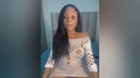 Media: Video of a young Black woman with long, straight black hair, wearing an off-shoulder, sheer white lace dress with cut-out designs, sitting on a bed with a beige headboard in a minimalistic bedroom.