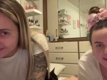 Media: Video of two women in a bathroom, one with blonde hair and tattoos, the other with brown hair in a pink headband. The room has a white vanity and shelves with toiletries.